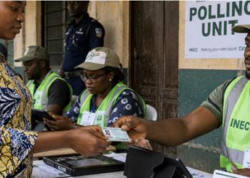 Close-up documentary-style photograph of a Nigerian voter presenting a Permanent Voter's Card to an INEC election official at a polling unit. Do not reproduce sensitive personal information on the card.